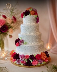 Luxury four-tier white wedding cake with floral embossing and colorful flower decorations.